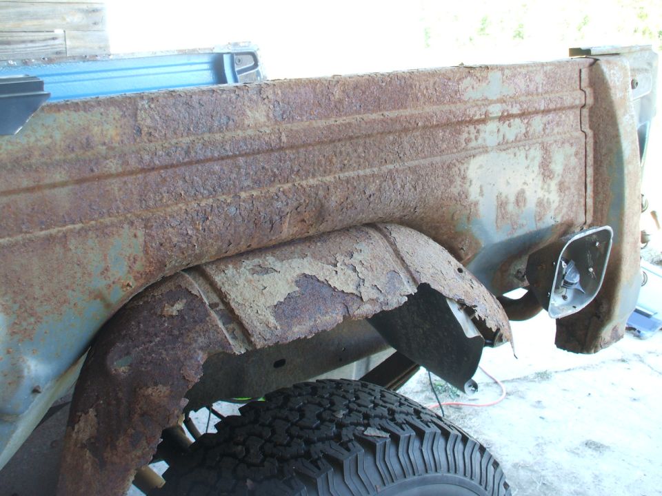 Heavily rusted rear passenger side of a truck bed and wheel well, showing significant body damage and peeling paint. A tire is visible below