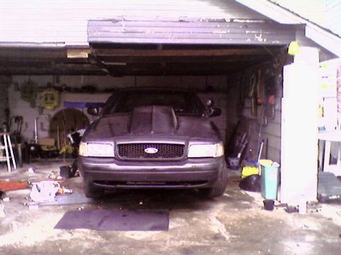 1998 Ford Crown Victoria - A dark sedan with a large hood scoop is parked inside a cluttered garage.