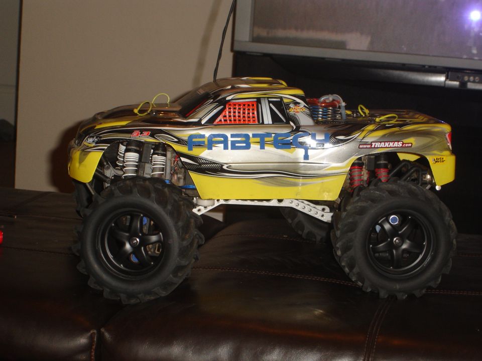 Yellow and silver remote control monster truck with "FABTECH" and "TRAXXAS" decals, on a brown leather surface