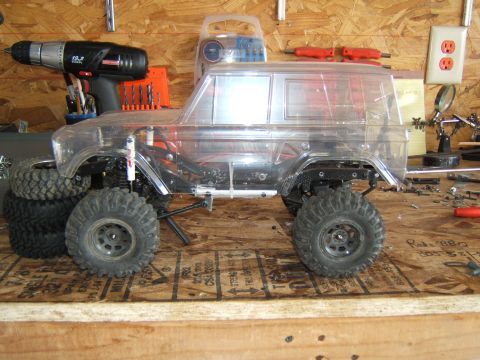 1988 Ford Bronco - RC crawler model with clear body sits on a workbench next to tools and spare tires.