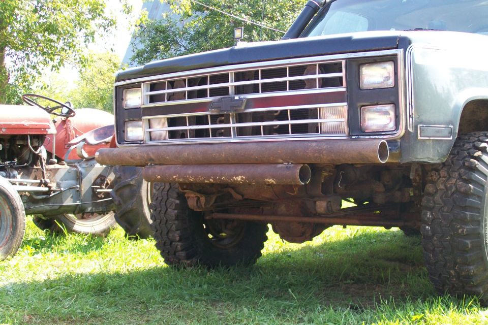 Front end of a dark SUV with a chrome grille and a rusty pipe bumper. A red tractor is parked next to it