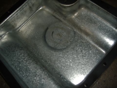 1988 Ford Bronco - Close-up looking down into the sump of a bare stamped-steel oil or transmission pan, showing the raw zinc-galvanized speckled finish, a cir…