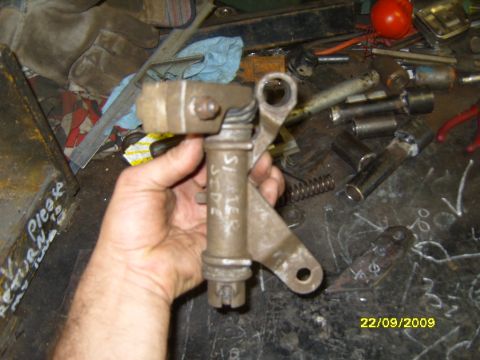 1989 Ford Bronco - A metal linkage part is held up for inspection, with various tools and metal pieces scattered on the workbench below.