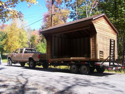 1979 Ford Bronco - A brown pickup truck tows a large wooden shed on a flatbed trailer down a paved road.