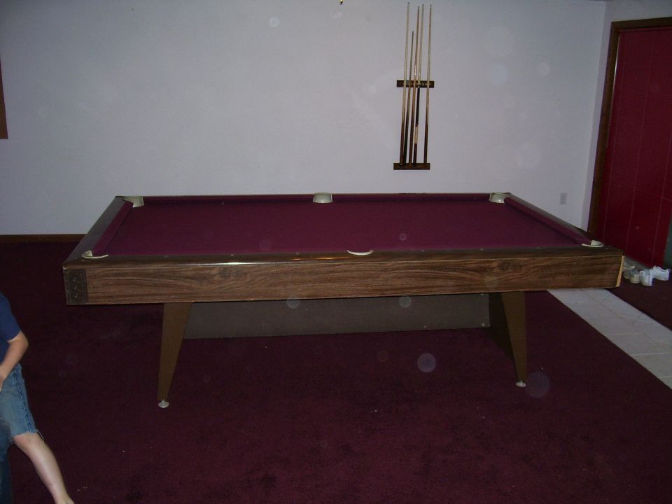 Pool table with maroon felt and wooden frame, cue rack on wall