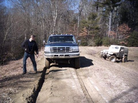 1989 Ford F-150 - Two off-road vehicles are stuck in a muddy rut on a dirt path surrounded by trees.