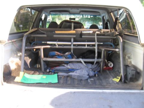 1984 Ford Bronco - The rear cargo area of a white SUV is filled with camping equipment and a metal roll cage.