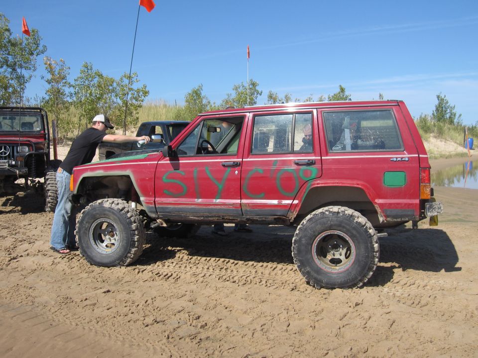 Lifted red Jeep Cherokee on big tires parked on sand with graffiti on doors