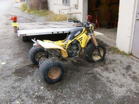 2000 Ford E350 - A yellow Yamaha three-wheeled ATV sits on a gravel lot next to a flatbed trailer.
