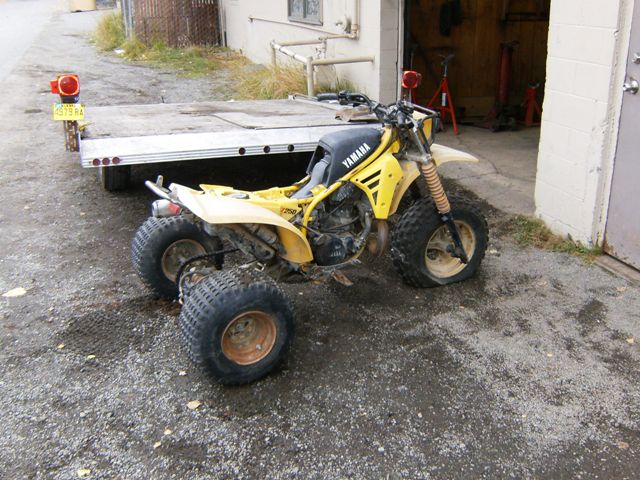 Yellow Yamaha three-wheeler parked on gravel next to a trailer