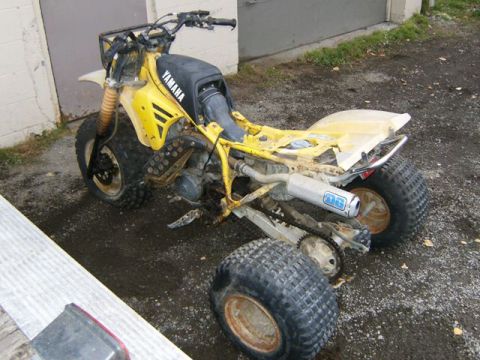 2000 Ford E350 - A yellow Yamaha three-wheeled ATV sits in a dirt lot, showing signs of disrepair and missing parts.