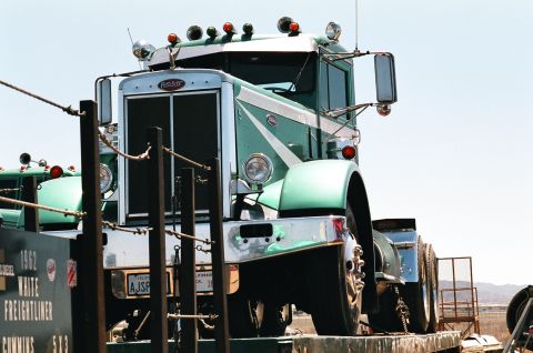 1981 Kenworth W900A - A green and white Peterbilt truck is seen from a 3/4 front angle. Another truck is partially visible to the left.