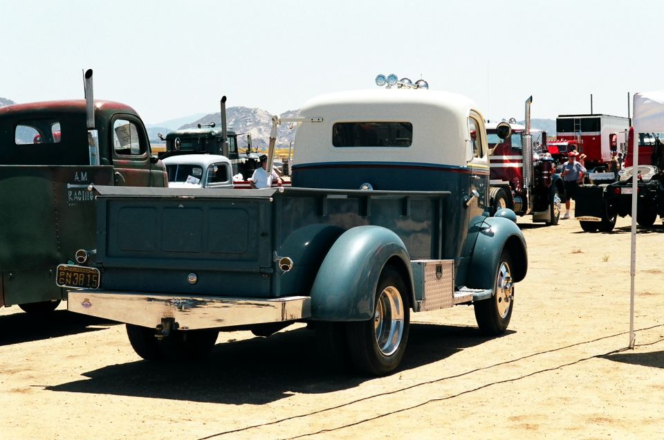 Rear view of a blue and white pickup truck with chrome bumper and dual exhaust stacks. Other trucks are parked nearby