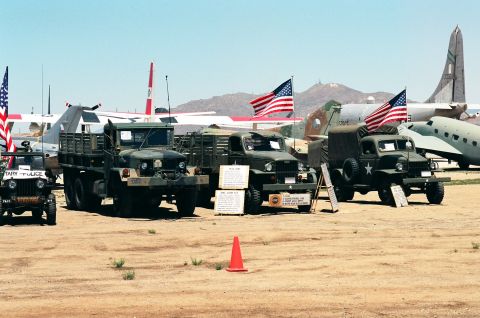 1981 Kenworth W900A - Several military vehicles and aircraft are parked outdoors on a sunny day, with American flags displayed.