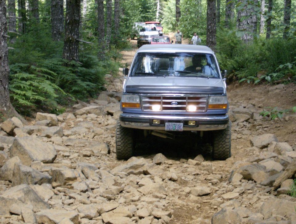 1996 Ford Bronco driving on a rocky, wooded trail with other vehicles behind it