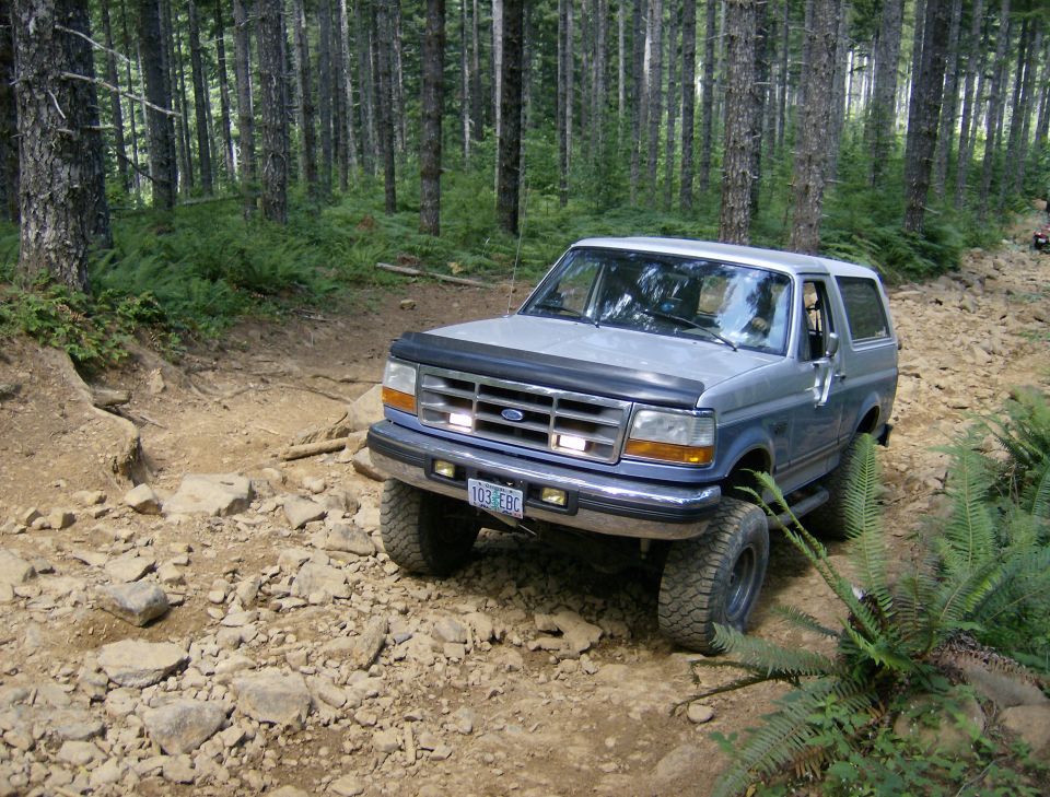 blue and silver 1996 Ford Bronco drives on a rocky, wooded trail
