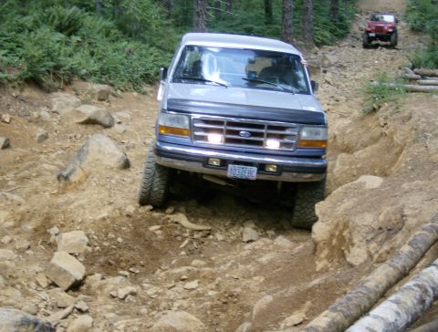 1996 Ford Bronco - A silver Ford Bronco drives off-road on a challenging, rocky path with another vehicle visible in the background.