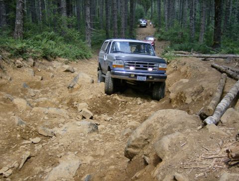 1996 Ford Bronco - A 1996 Ford Bronco navigates a challenging rocky path through a dense forest, with other vehicles visible further up the trail.