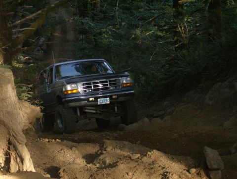 1996 Ford Bronco - A dark blue Ford Bronco navigates a challenging off-road trail through a wooded area.