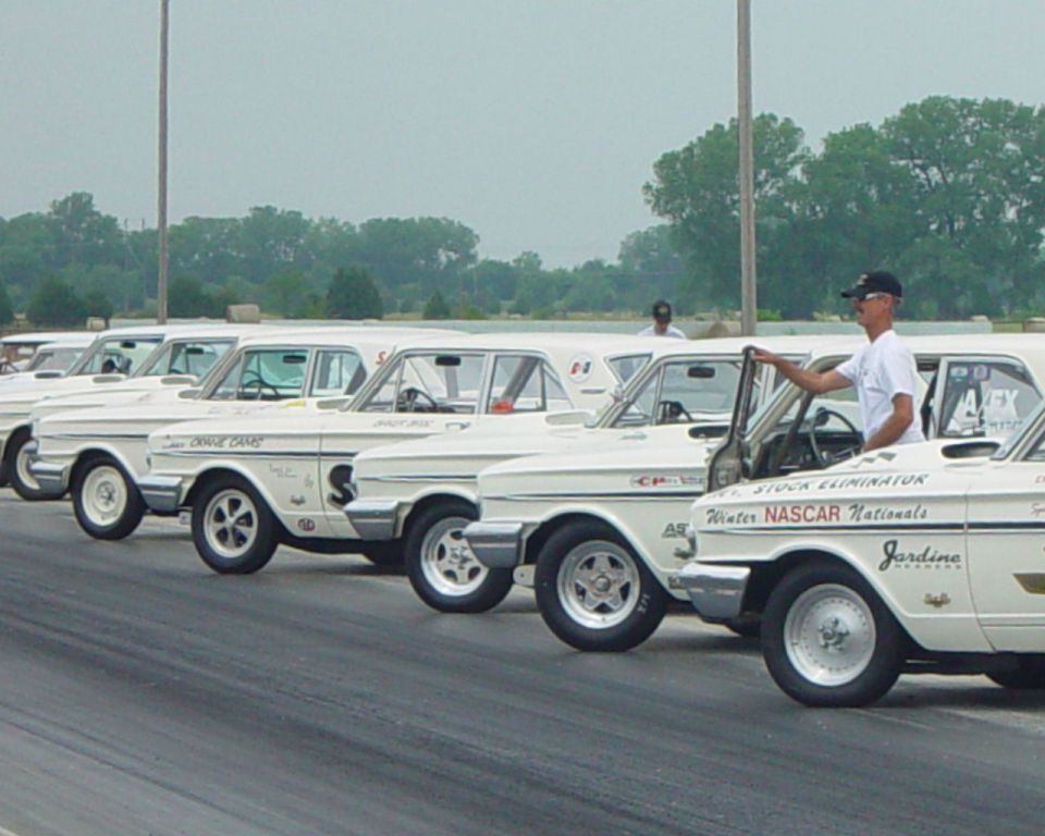 Row of white 1960s lightweight drag cars with bubble hoods staged at a dragstrip