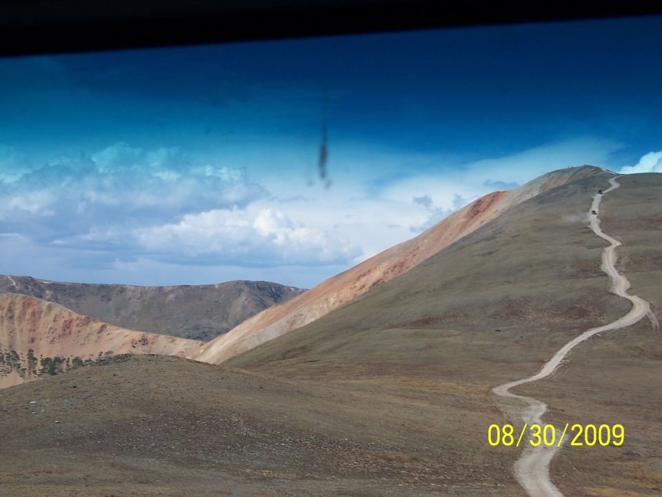 Several vehicles drive up a winding dirt road on a mountain under a cloudy blue sky