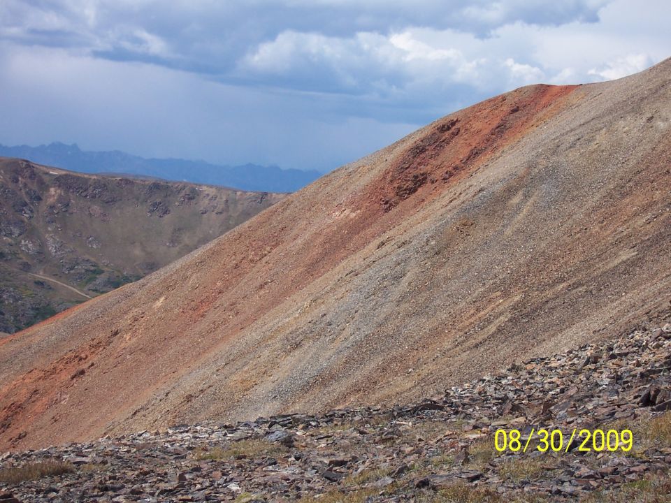Mountain slope with red and gray rocks under a cloudy sky. A dirt road is visible in the distance