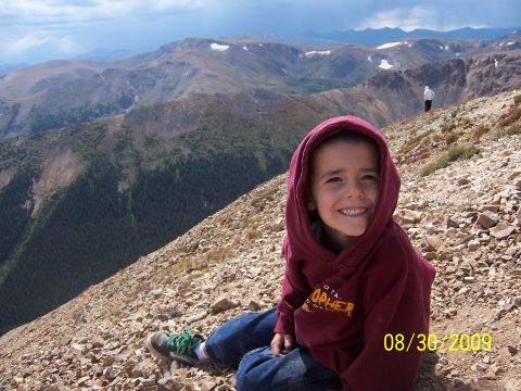 1994 Ford Bronco - A young child smiles while sitting on a rocky mountain slope, with a panoramic view of distant mountains under a cloudy sky.