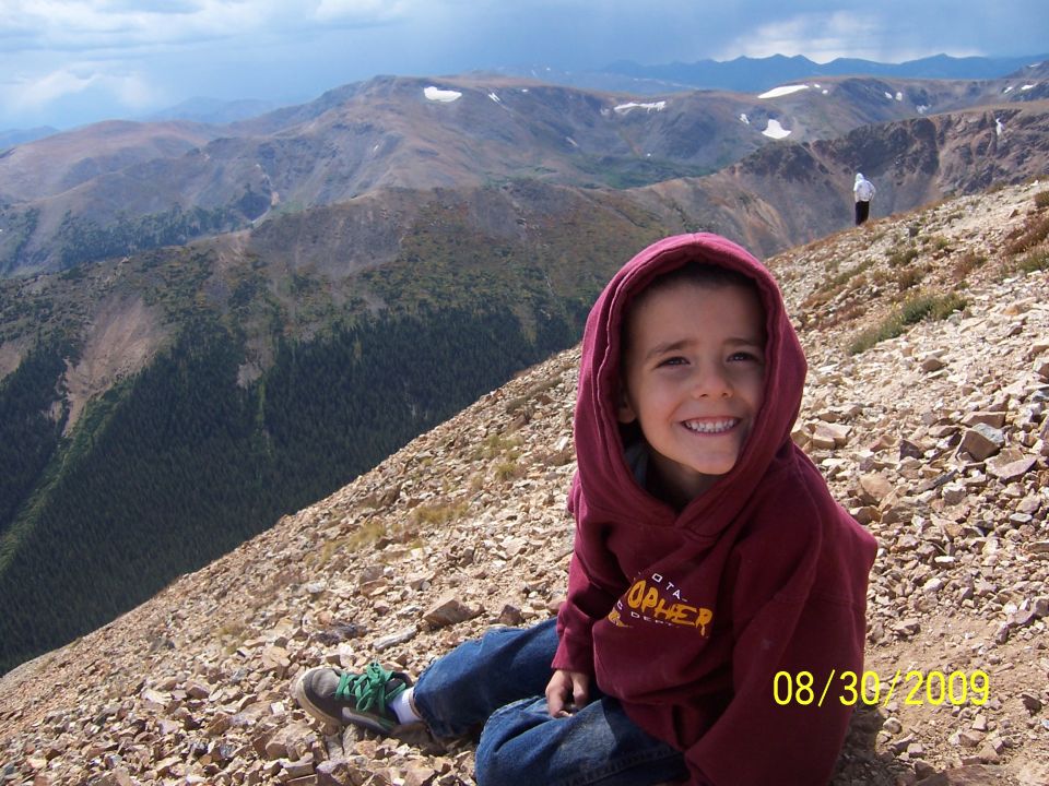 Child in maroon hoodie and jeans sits on a rocky mountain slope with a vast mountain range in the background
