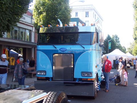1981 Kenworth W900A - A blue Peterbilt truck is parked on a street, with onlookers and tents in the background.