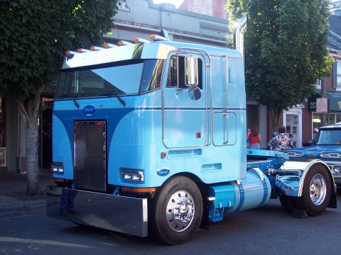 1981 Kenworth W900A - A blue Peterbilt semi-truck cab is parked on a street, with another truck visible in the background.