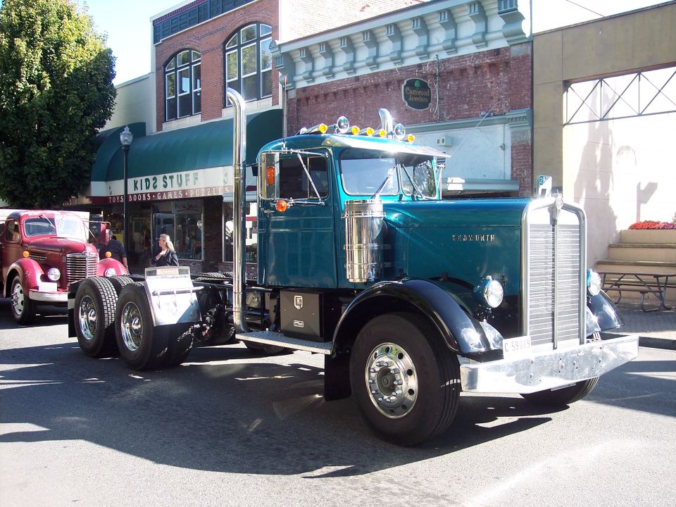 Teal Kenworth truck with chrome stacks and grille, parked on street. Another red truck is visible