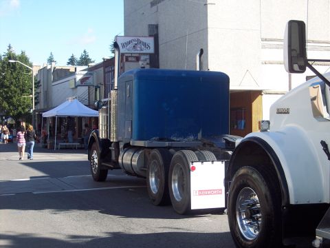 1981 Kenworth W900A - Two Kenworth trucks are parked on a street with buildings and tents visible in the background.