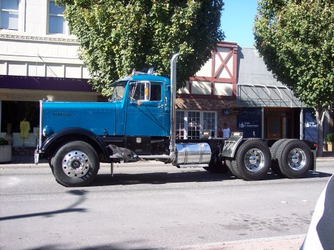 1981 Kenworth W900A - A blue 1981 Kenworth W900A semi-truck is parked on a street with buildings and trees in the background.