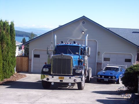 1981 Kenworth W900A - A blue 1981 Kenworth W900A semi-truck is parked in a driveway next to a blue sports car, with a house and scenic background.
