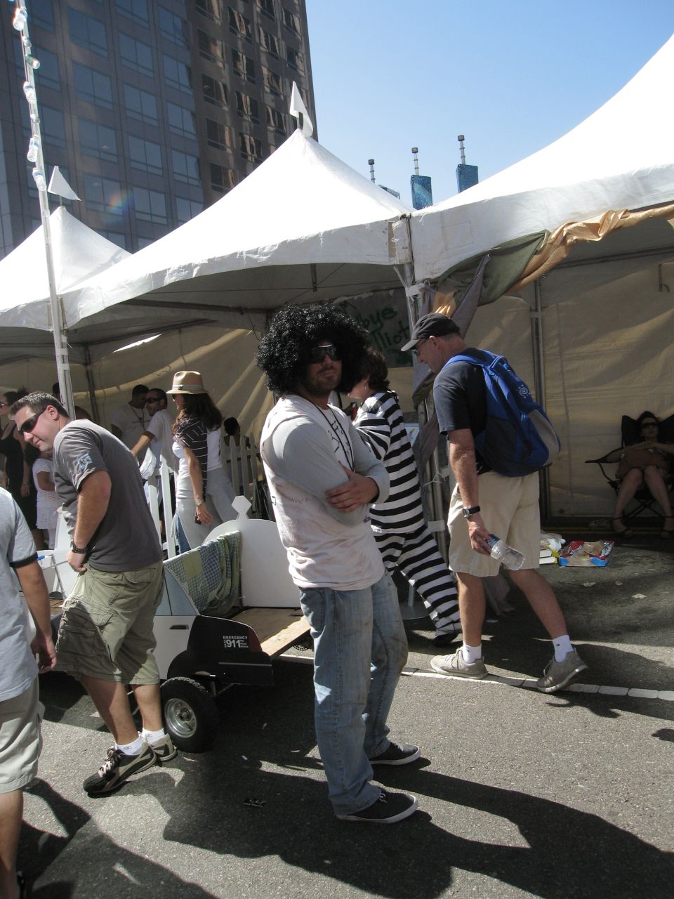 People in casual clothes and costumes stand around tents at an outdoor event. One person wears a large afro wig and sunglasses