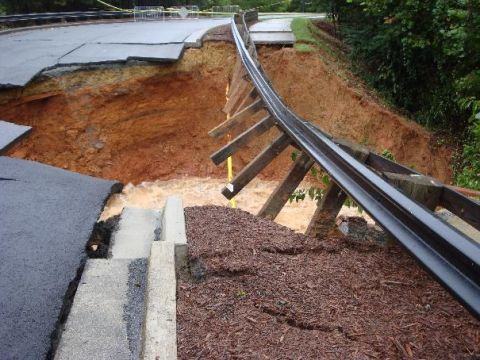 1979 Ford F-150 - A section of road has collapsed due to flooding, revealing a large chasm with rushing water below and a damaged guardrail.