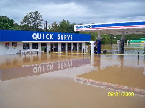 1979 Ford F-150 - A gas station and convenience store are submerged in floodwaters, with "Quick Serve" and "Marathon" signage visible above the water line.