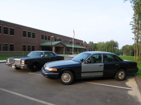 2001 Ford Crown Victoria - A two-tone police car, likely a 2001 Ford Crown Victoria, is parked in a lot with another older vehicle in front of a school.