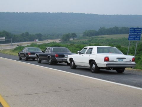 2001 Ford Crown Victoria - Three Ford Crown Victorias drive in a line on a highway with a scenic, hilly background.
