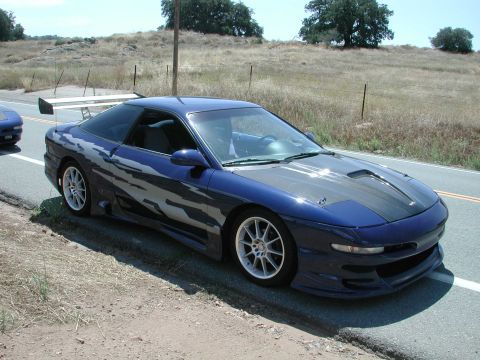 1990 Ford Probe - A modified 1990 Ford Probe with custom graphics and a large rear spoiler sits parked on the side of a road.