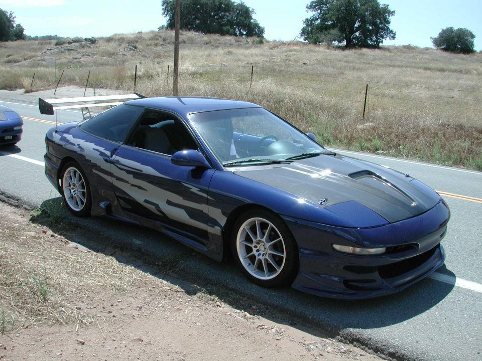 Dark blue modified coupe with rear wing and white wheels on a rural roadside