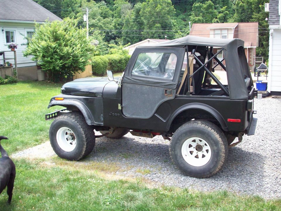 Black Jeep CJ5 side view with soft top and white wheels on gravel driveway