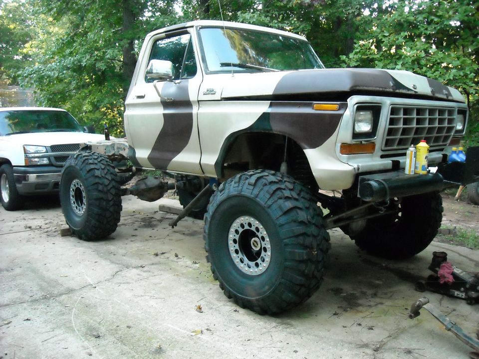 1979 Ford F-150 pickup truck with camouflage paint and large off-road tires, parked next to a white Chevrolet pickup