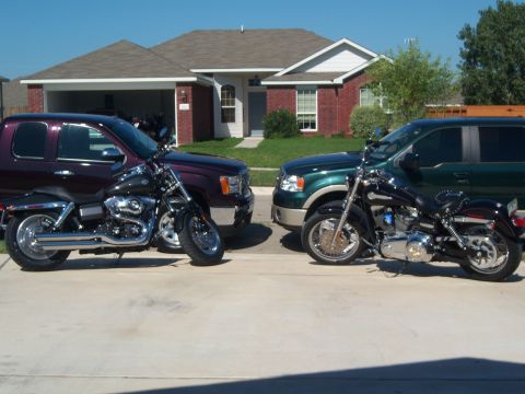 1996 Ford Bronco - Two motorcycles are parked in a driveway between a dark purple SUV and a green pickup truck.