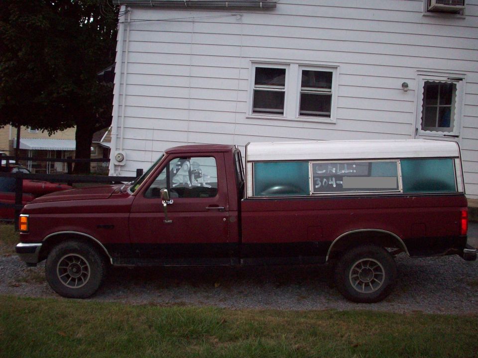 Maroon Ford pickup with white camper shell parked on gravel, 'For Sale' and phone number signs in windows.