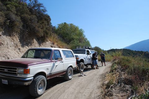 1996 Ford Bronco - Two Ford Broncos, one with a maroon top, are parked on a dirt road with hills and trees in the background.