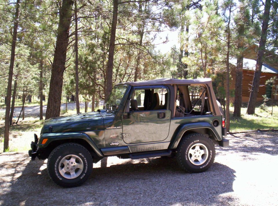 Green 2002 Jeep TJ Wrangler parked outdoors with trees and a cabin in the background