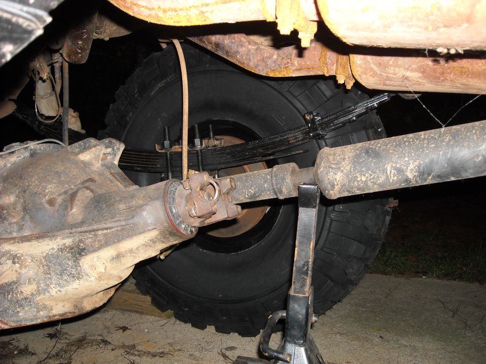 Close-up of a vehicle's rear axle, leaf springs, driveshaft, and tire, supported by a jack stand