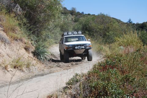 1996 Ford Bronco - A white SUV navigates a dusty, off-road trail surrounded by vegetation.