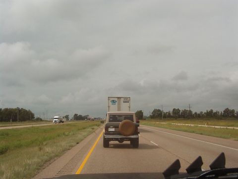 1975 Ford Bronco - A Ford Bronco is seen from behind on a highway, towing a large trailer under a cloudy sky.